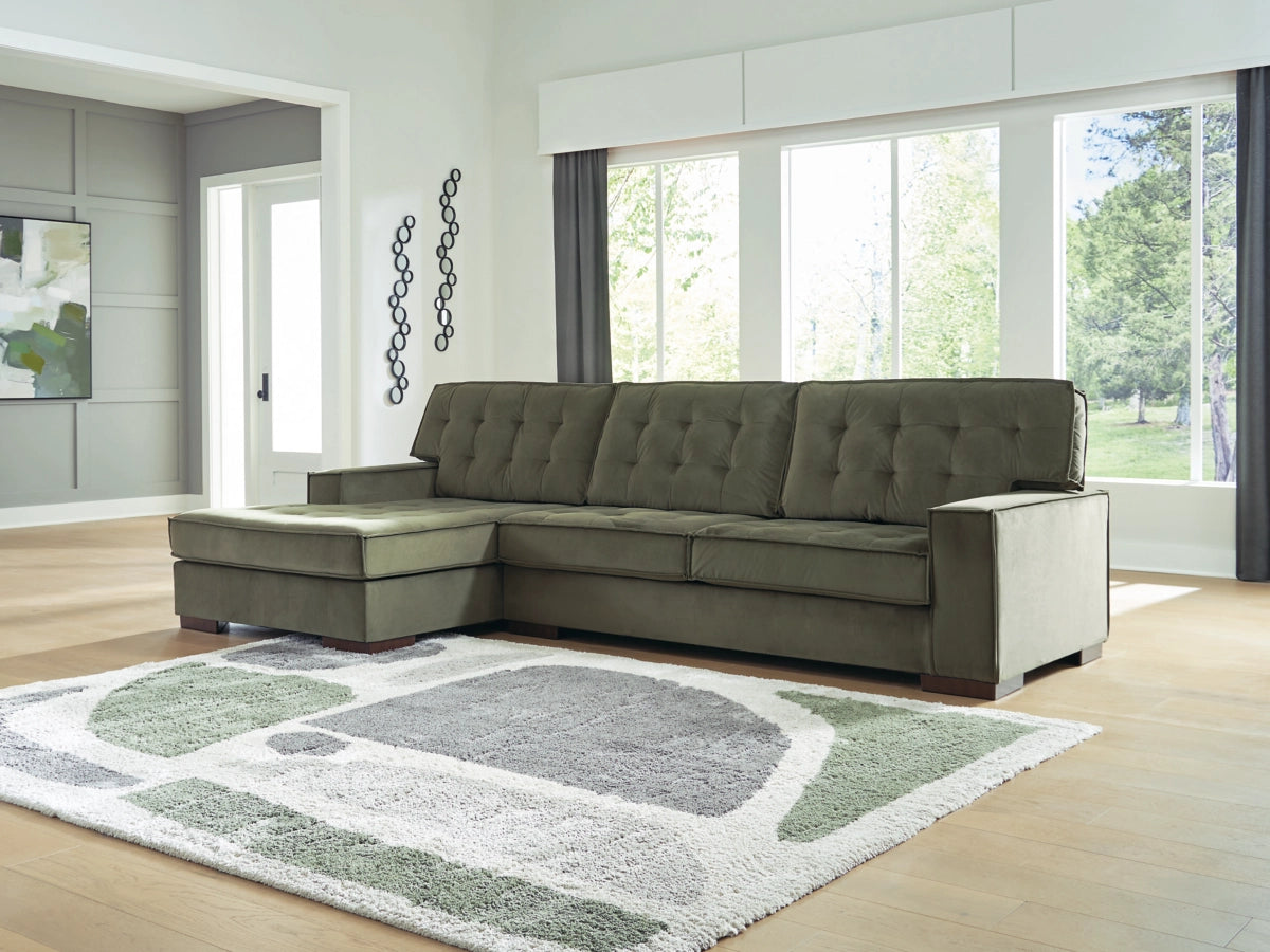 Green sectional sofa in a living room with large windows and a patterned rug.
