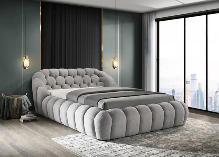 Gray tufted bed in a modern bedroom with large windows and cityscape view.