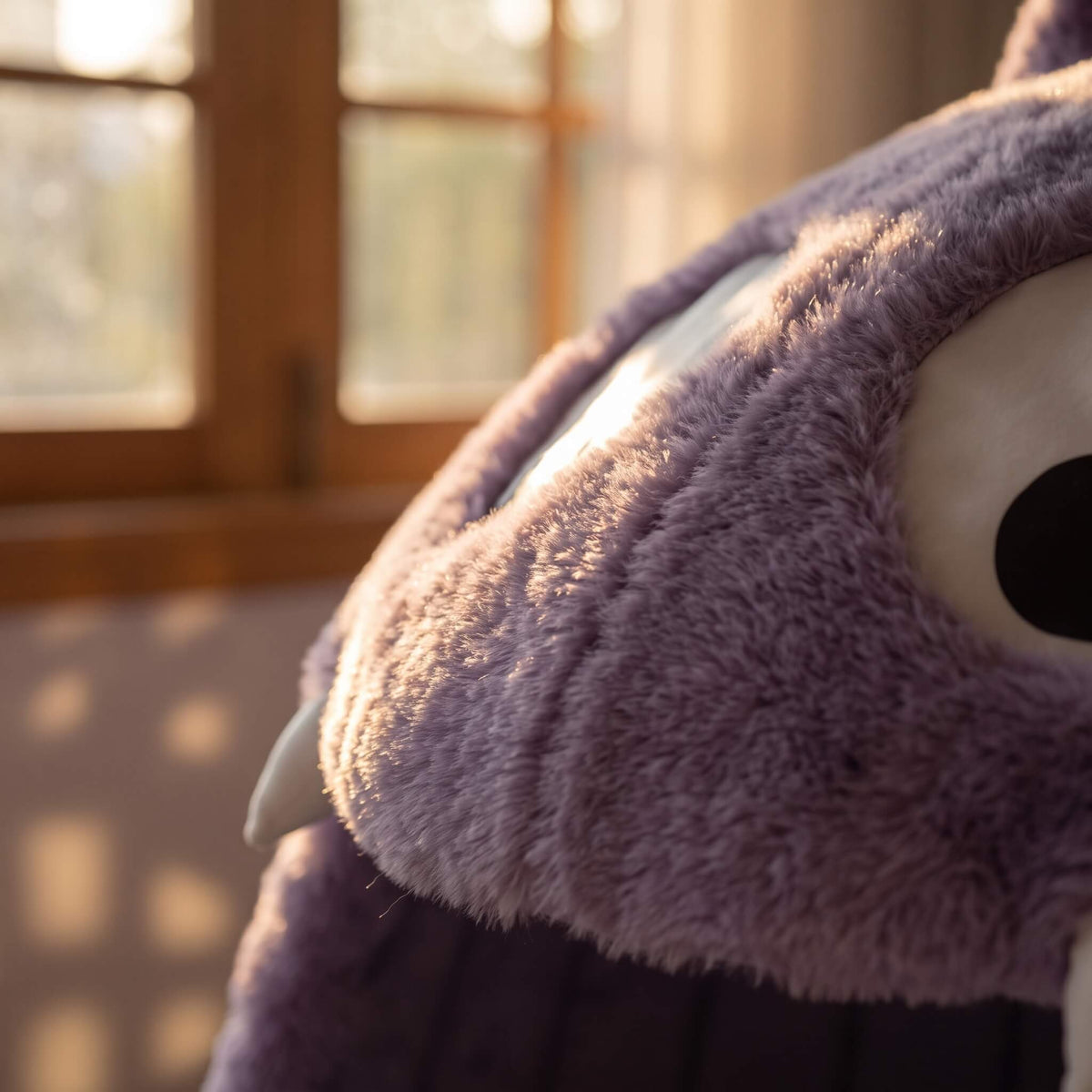 A close-up shot of a purple plush toy with soft, fuzzy texture, featuring a large white eye with a black pupil and a small white tooth, set against a warm, softly blurred background with a wooden window frame.