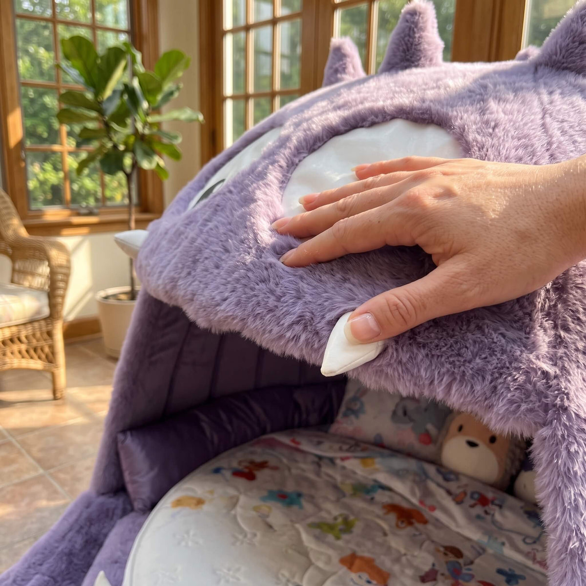 A close-up of a person's hand touching the plush purple hood of a pet bed, which features a cute monster face design. The bed is set in a cozy, sunlit room with a window, potted plant, and wicker chair visible in the background.