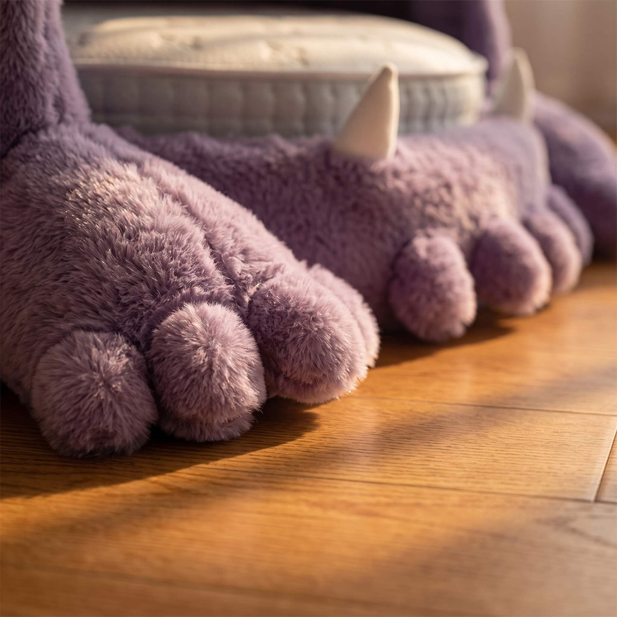 A close-up of plush monster-style slippers in lavender purple with small white horns, photographed on a wooden floor in warm sunlight.