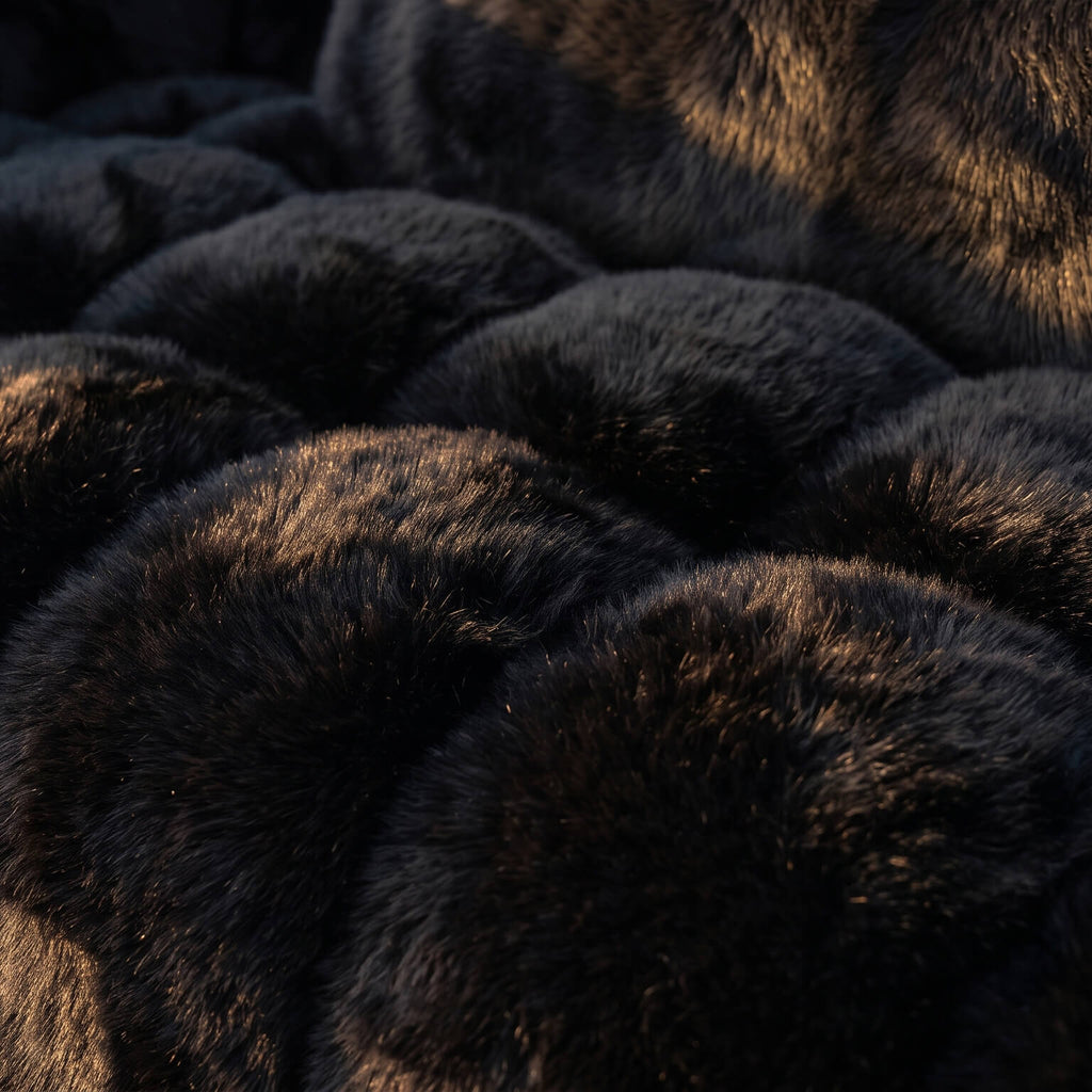 A close-up of dark faux fur fabric with a textured, scalloped pattern, illuminated by warm golden light against deep shadows.