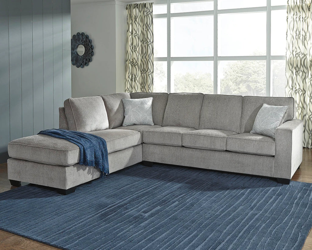 Light gray L-shaped sectional sofa with chaise lounge in a modern living room featuring a blue area rug, large windows, and textured accent pillows.