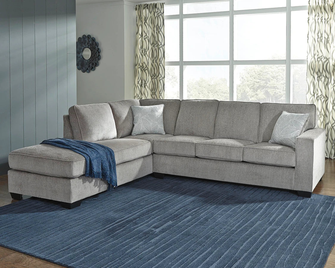 Light gray L-shaped sectional sofa with chaise lounge in a modern living room featuring a blue area rug, large windows, and textured accent pillows.
