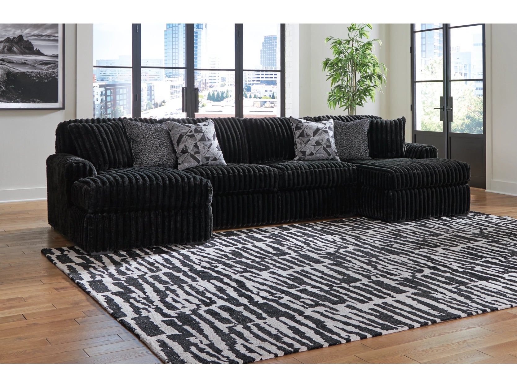 Black sectional sofa with patterned cushions on a black and white rug in a room with large windows.