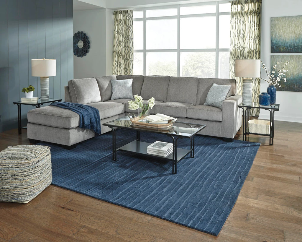 Contemporary gray fabric sectional couch with chaise in a staged living room with blue rug, glass coffee table, wood flooring, and decorative lighting.